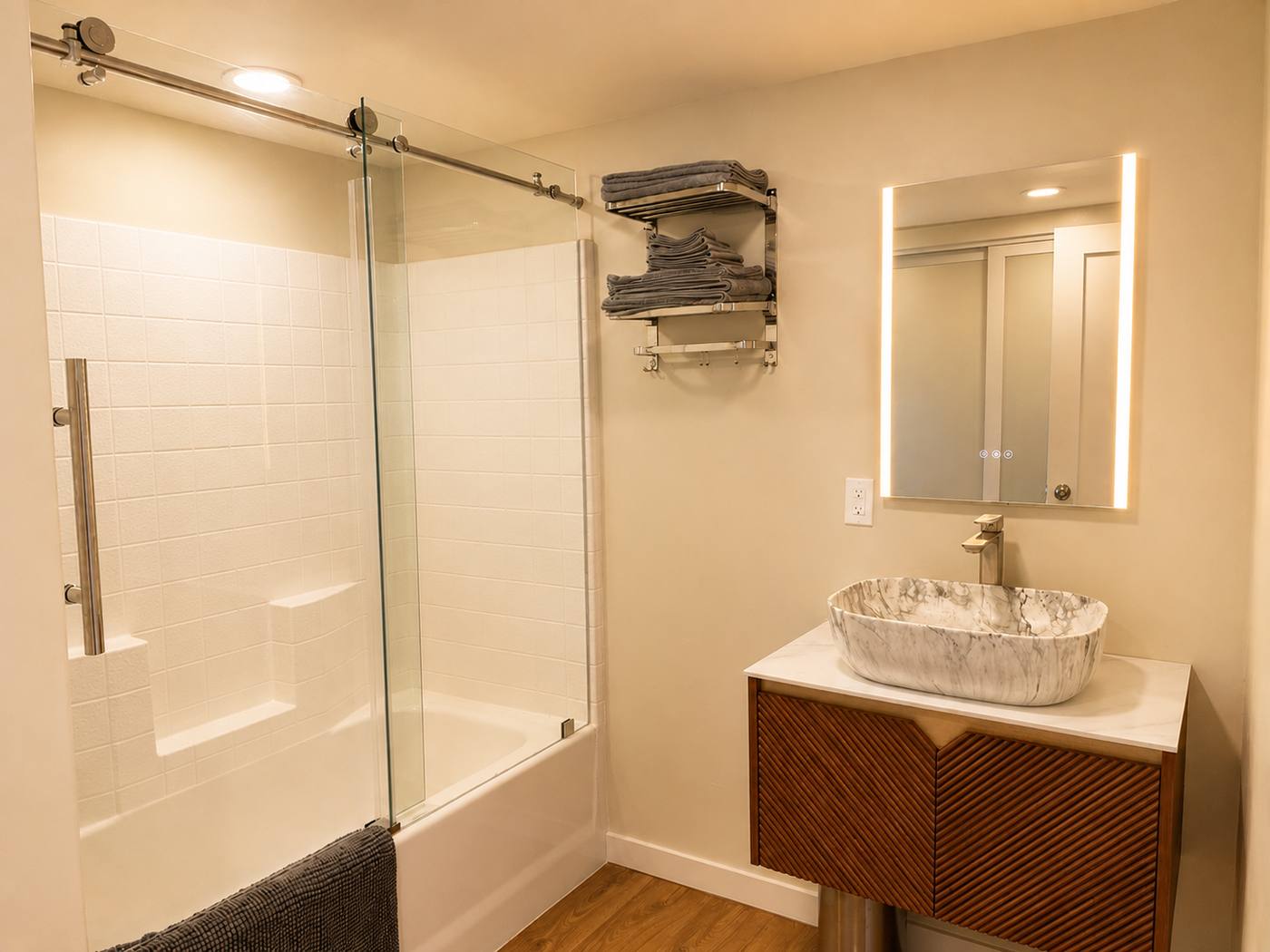 Bathroom with a marble vessel sink on a fluted walnut vanity, backlit mirror and glass-door shower over tub
