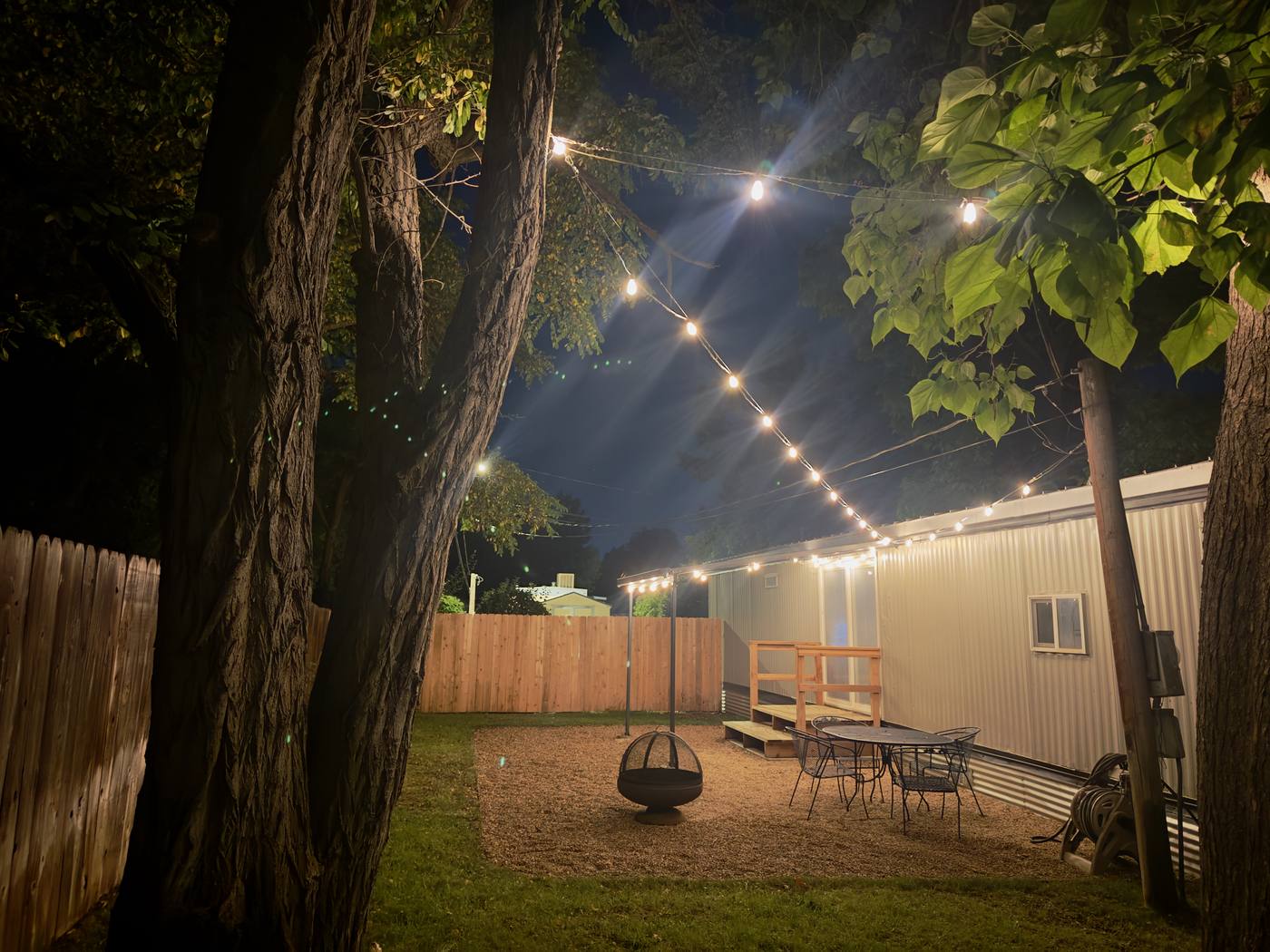 The back yard after dark, string lights strung between the trees above the fire pit